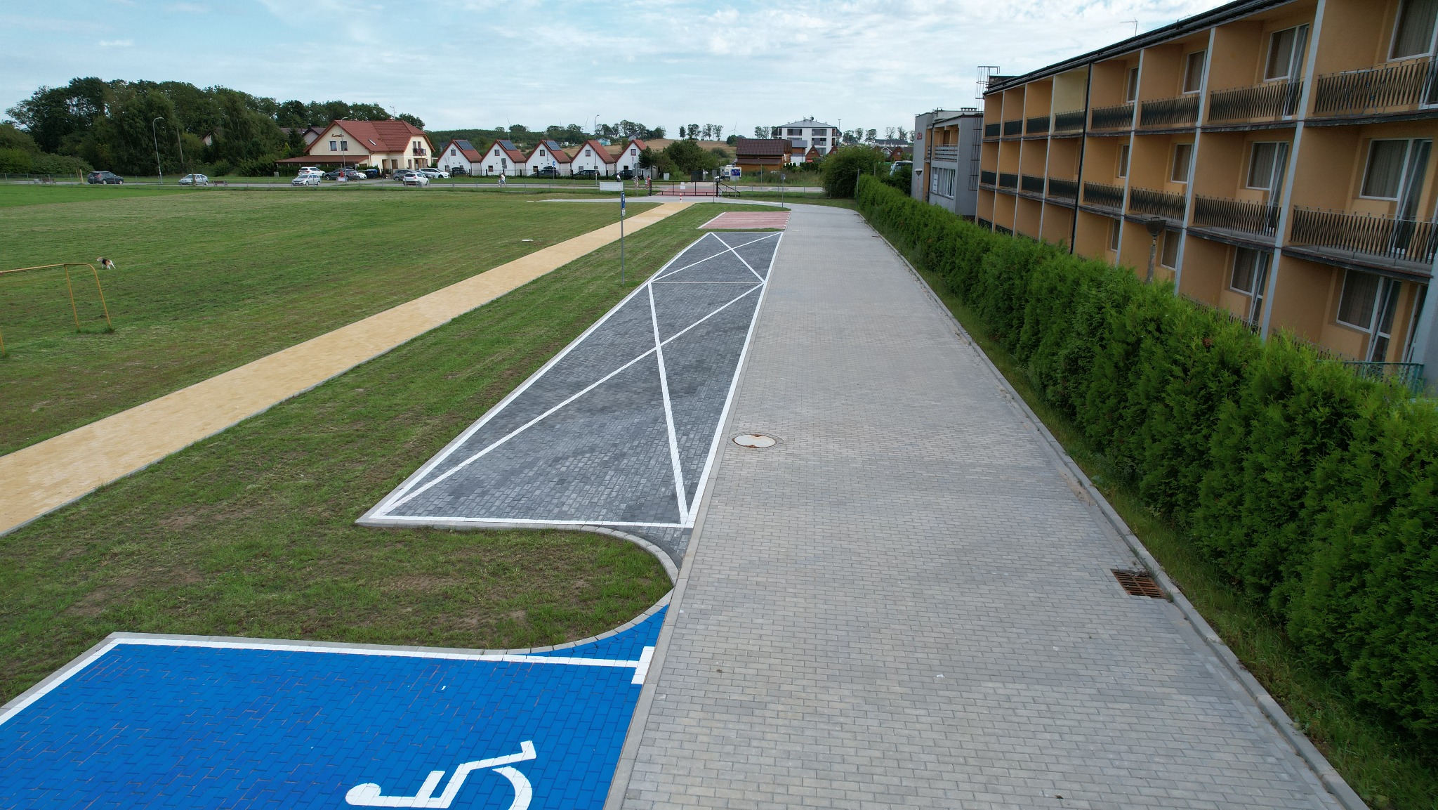 Nowa nawierzchnia z kostki brukowej: parking z wyznaczonym miejscem dla niepełnosprawnych, ścieżka z jasnego kruszywa, budynek w tle, widok z góry.