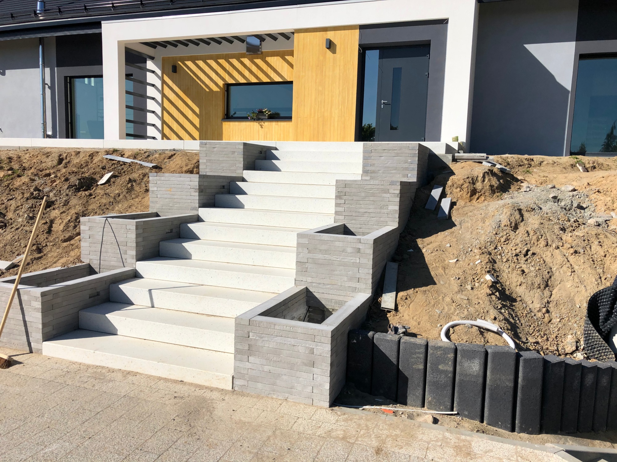 Nowoczesne, białe schody wejściowe z szarej kostki, prowadzące do domu z żółtą elewacją, widoczne przygotowane podłoże i elementy obrzeża.