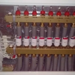 Rozdzielacz ogrzewania podłogowego w szafce, z widocznymi zaworami termostatycznymi, rurami i modułem sterującym z przewodami.