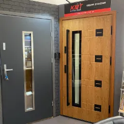 Ekspozycja dwóch modeli drzwi wejściowych: szare drzwi z wąskim przeszkleniem i srebrną klamką oraz drzwi w odcieniu drewna z czarnymi akcentami i długim uchwytem, obok stojak z katalogami.