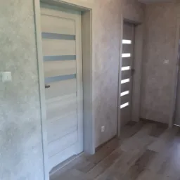 Białe drzwi wewnętrzne z poziomymi przeszkleniami w jasnym korytarzu z drewnianą podłogą, widoczne gniazdko elektryczne na ścianie.