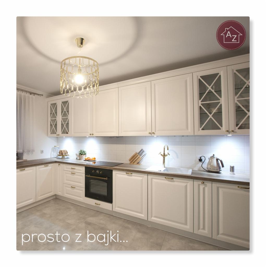 Jasna kuchnia z białymi szafkami, złotymi uchwytami i ozdobnym, złotym żyrandolem, wykończona białymi płytkami i szarą podłogą, z delikatnym oświetleniem podszafkowym.