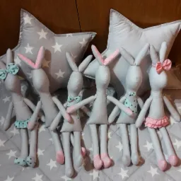 Ręcznie szyte szare króliczki z różowymi i turkusowymi akcentami, ustawione na kocu w gwiazdki, z poduszkami w kształcie gwiazd w tle.