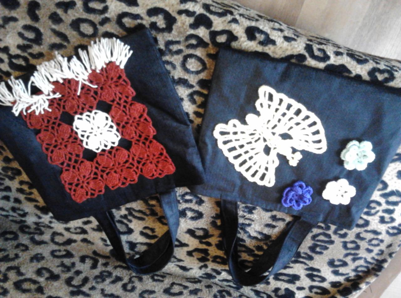 Dwie ciemne torby na zakupy z ręcznie wykonanymi, szydełkowymi aplikacjami: czerwono-białą kwadratową z frędzlami i białą w kształcie anioła z trzema kwiatkami, leżące na tkaninie w panterkę.