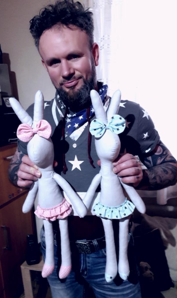 Osoba prezentuje dwa ręcznie wykonane, szmaciane króliki w sukienkach z kokardami, z tatuażami na rękach i w bluzie z motywem amerykańskim.