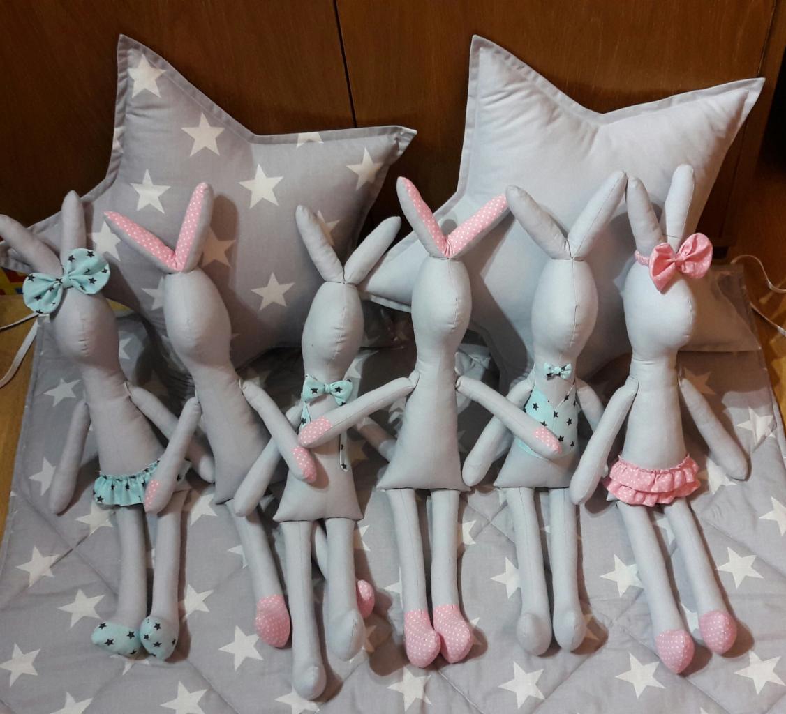 Ręcznie szyte szare króliczki z różowymi i turkusowymi akcentami, ustawione na kocu w gwiazdki, z poduszkami w kształcie gwiazd w tle.