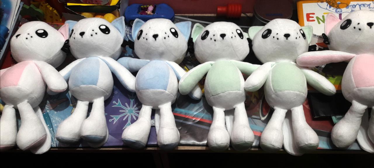 Rząd pluszowych zabawek w kształcie białych zwierzątek z pastelowymi akcentami (różowy, niebieski, zielony), ustawionych na tle zabawek i książek.