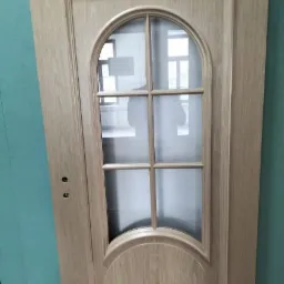 Jasne, drewniane drzwi z zaokrąglonym, przeszklonym panelem, widoczne otwory na klamkę, na tle ściany w kolorze turkusowym.