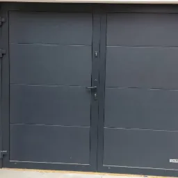 Grafitowa brama garażowa dwuskrzydłowa z poziomymi panelami, zamontowana w białej elewacji, obok widoczny fragment drzwi wejściowych z panelem kodowym.