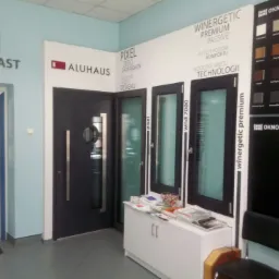 Ekspozycja w salonie: czarne drzwi wejściowe z okrągłym przeszkleniem i długim uchwytem, obok okna z czarną ramą i tablica z próbkami kolorów, całość prezentowana na tle ścian z logo producentów.