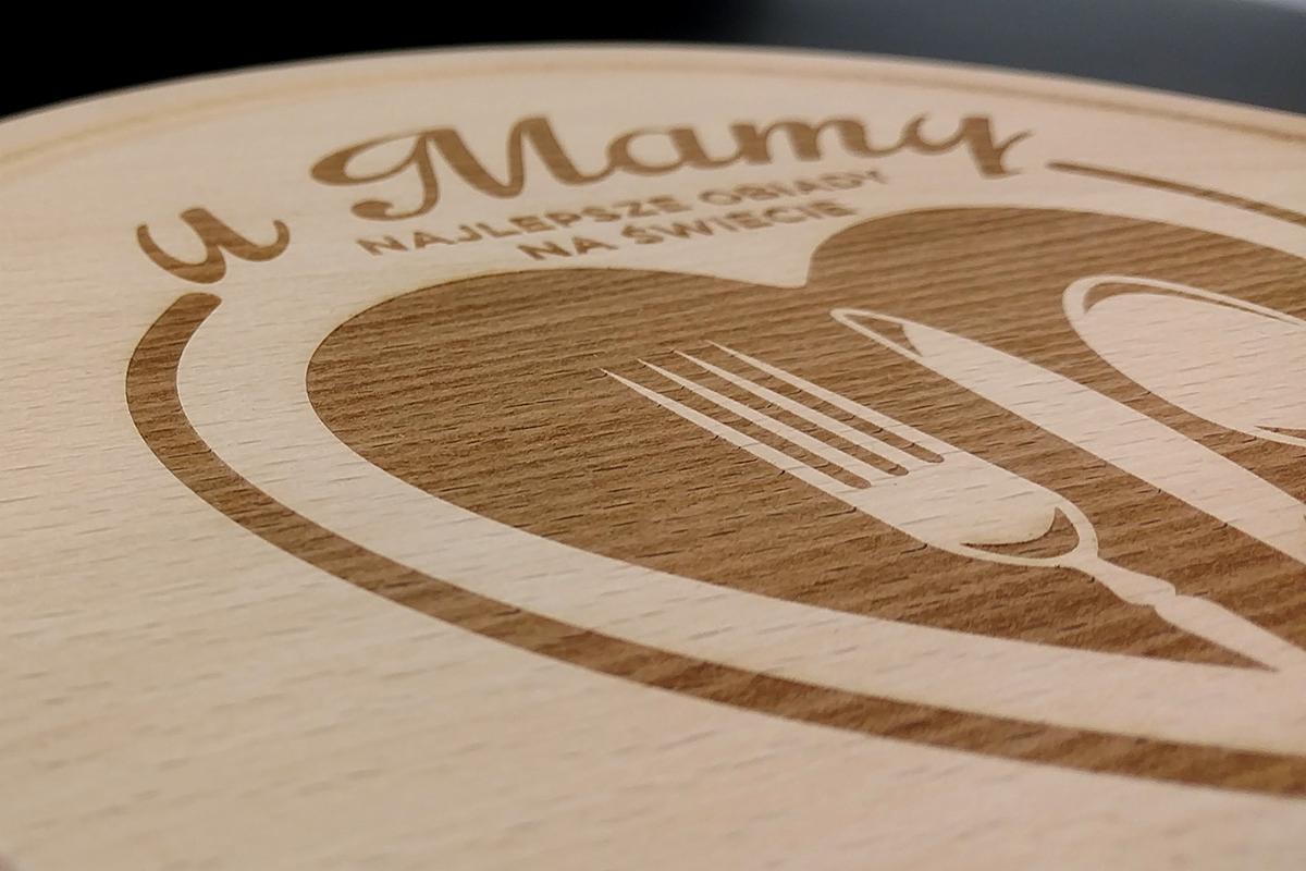 Drewniana deska z wygrawerowanym napisem 'U Mamy Najlepsze Obiady Na Świecie' oraz grafiką przedstawiającą talerz, nóż i widelec.