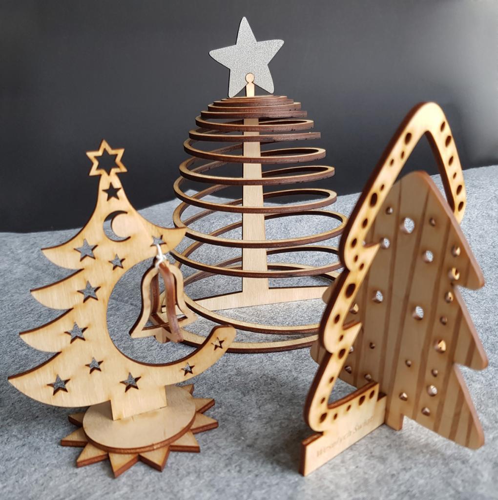 Trzy drewniane choinki dekoracyjne, każda w innym stylu: ażurowa z gwiazdami i księżycem, spiralna ze srebrną gwiazdą na szczycie, oraz pełna z dziurkami, stojące na szarym tle.