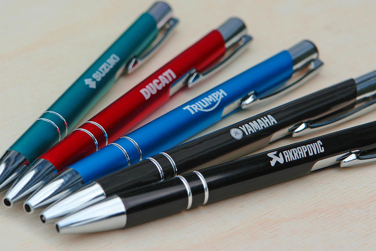 Pięć kolorowych długopisów z nadrukowanymi logotypami firm motoryzacyjnych: Suzuki, Ducati, Triumph, Yamaha i Akrapovic, leżące ukośnie na jasnym, drewnianym blacie.
