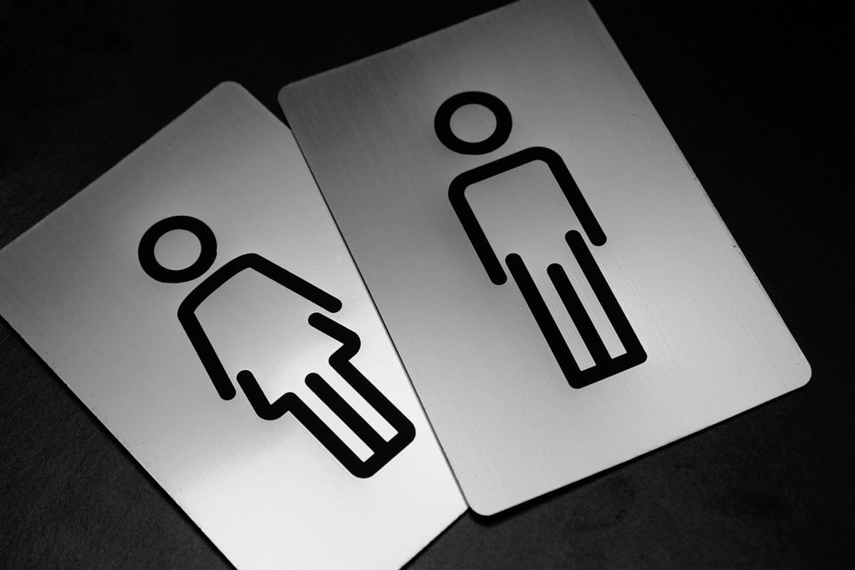 Dwie metalowe tabliczki z piktogramami toalet męskiej i żeńskiej, na czarnym tle, ukazujące minimalistyczne sylwetki.