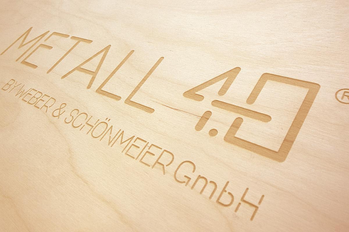 Jasne drewno z wygrawerowanym laserowo napisem 'METALL 4.0 BY WEBER & SCHÖNMEIER GmbH', widoczne delikatne usłojenie drewna.
