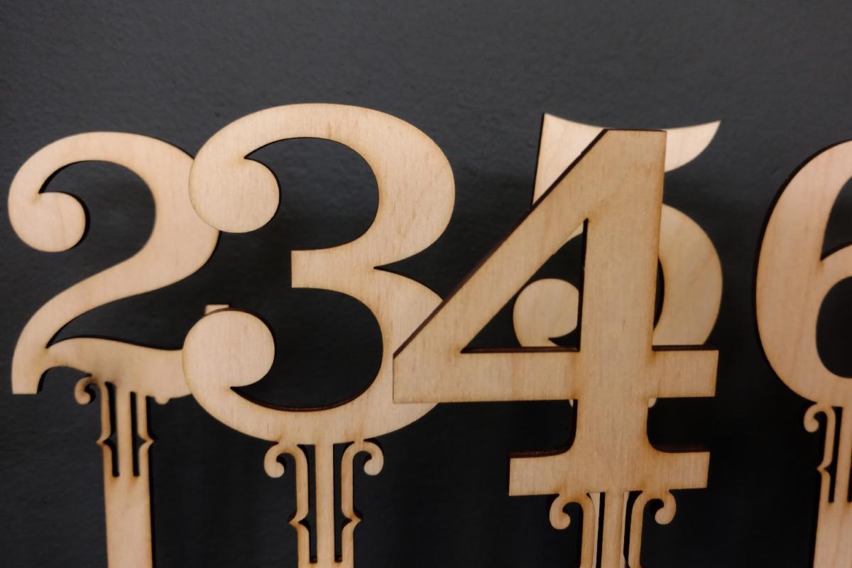 Wycięte laserowo z jasnego drewna numery 2, 3, 4, 5 i 6 z ozdobnymi podstawkami na ciemnoszarym tle.