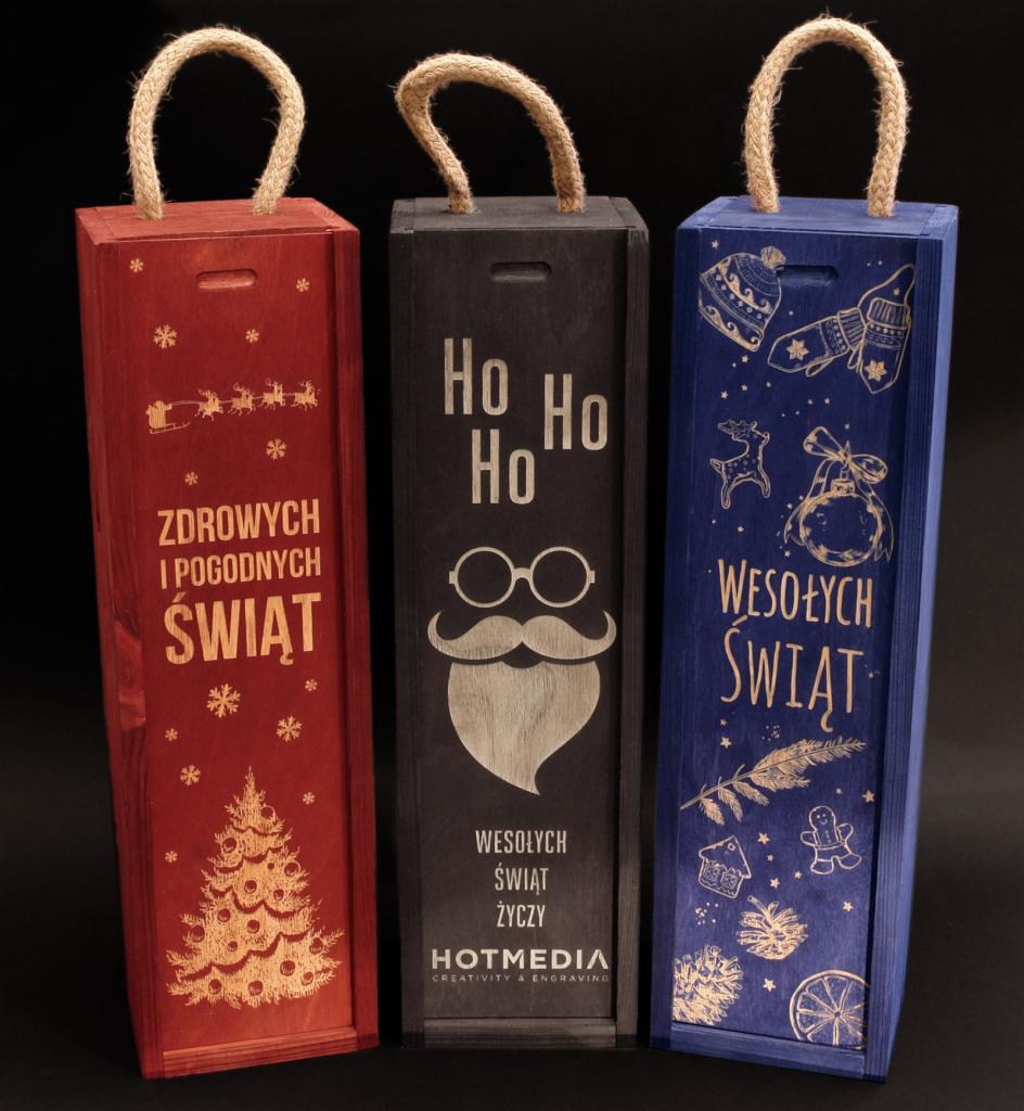Trzy drewniane pudełka prezentowe na wino w kolorach czerwonym, czarnym i niebieskim, z wygrawerowanymi świątecznymi motywami i tekstami, z uchwytami ze sznurka.