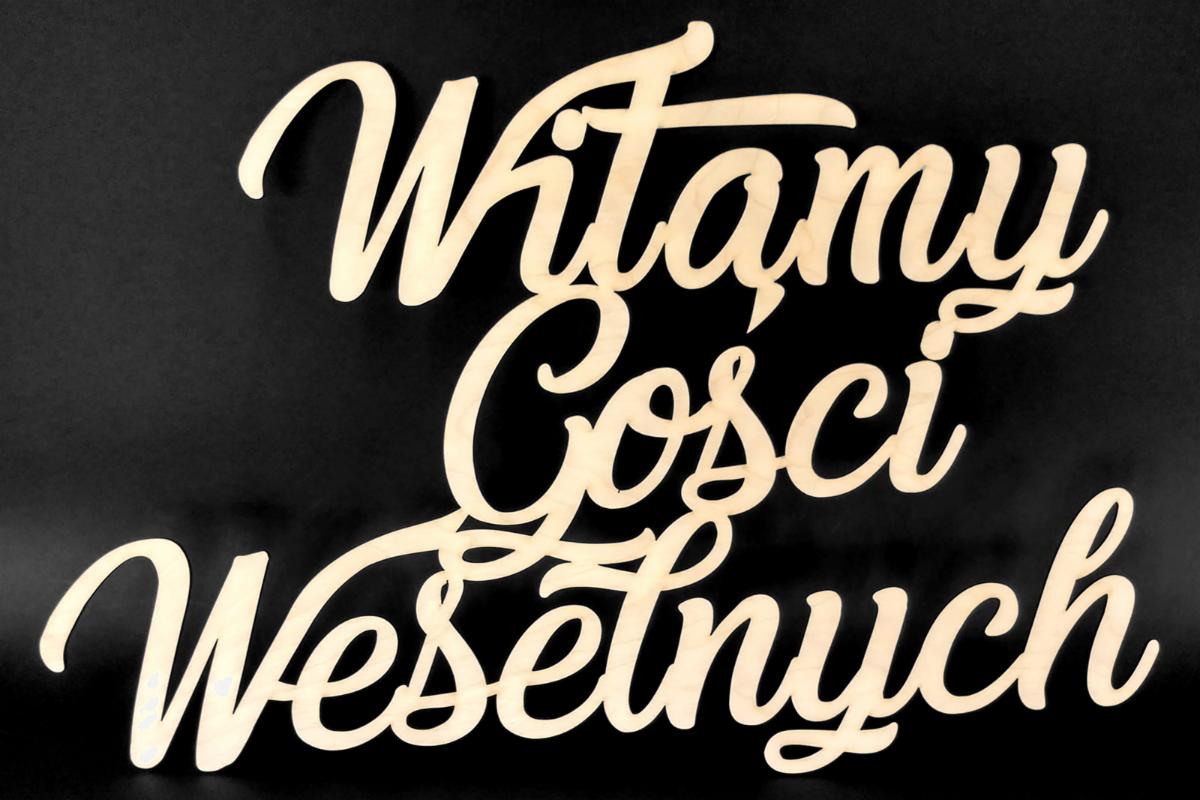 Drewniany napis 'Witamy Gości Weselnych' w ozdobnej czcionce na czarnym tle.