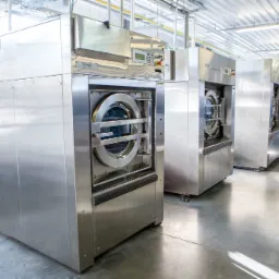 Przemysłowe pralki w nierdzewnej obudowie, ustawione rzędami w jasnym pomieszczeniu pralni, widoczne wózki z tekstyliami w tle.