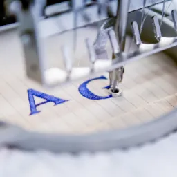 Detail haftu maszynowego: igła wyszywa niebieską literę 'C' na beżowym materiale w cienkie paski, widoczne elementy metalowej ramy i mechanizmu maszyny.