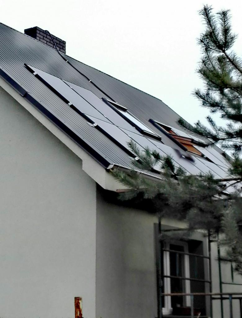 Panele słoneczne zamontowane na dachu domu z blachy trapezowej, widoczne okna dachowe i komin, z boku kadru gałąź sosny.