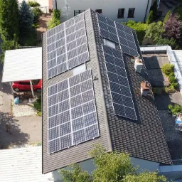 Widok z góry na dom z panelami słonecznymi na dachu, obok taras na dachu z roślinami, czerwony samochód w wiacie, pracownicy przy samochodzie dostawczym.