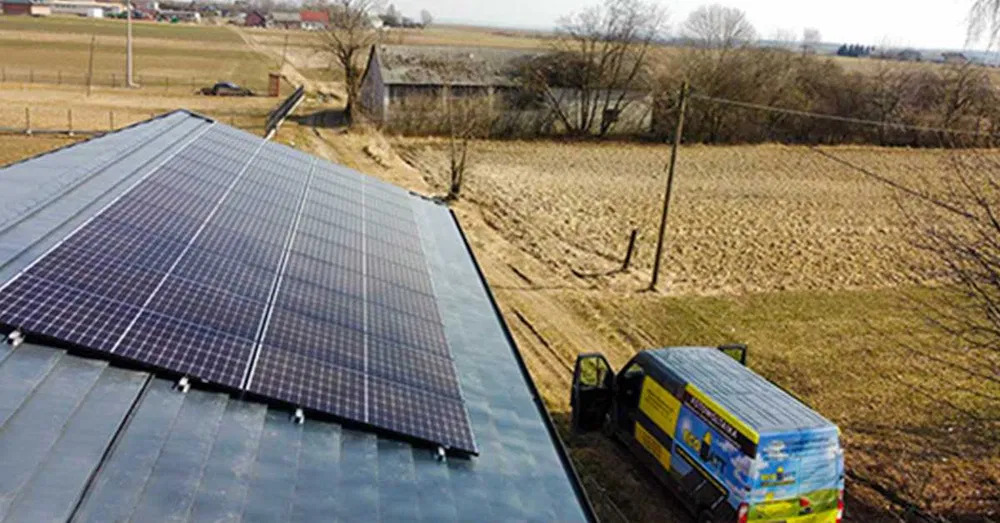 Montaż paneli fotowoltaicznych na dachu domu jednorodzinnego, widoczny firmowy bus zaparkowany obok, w tle pola uprawne i zabudowania wiejskie.