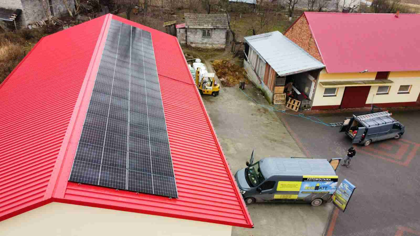 Widok z góry na czerwoną blachodachówkę z panelami, zaparkowane busy z logo firmy instalacyjnej oraz wózek widłowy w gospodarstwie.