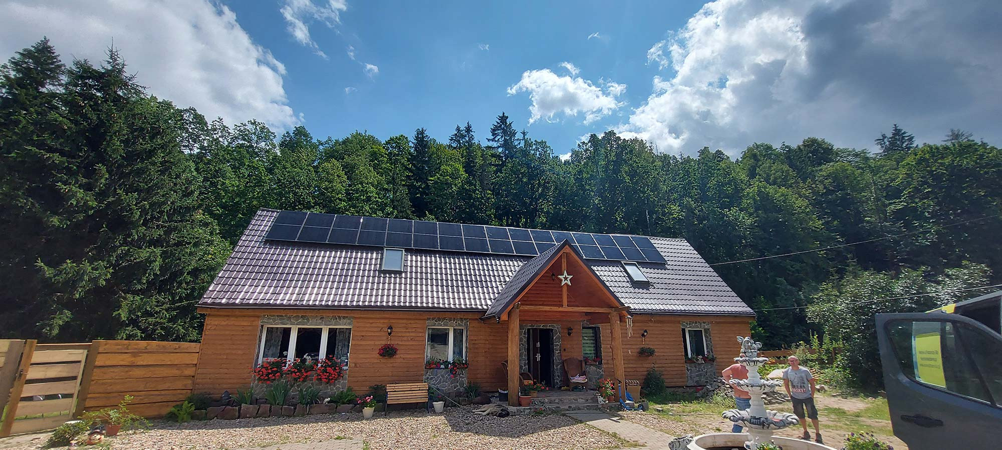 Drewniany dom z brązowym dachem, na którym zamontowano czarne panele słoneczne; w tle las i błękitne niebo z chmurami.
