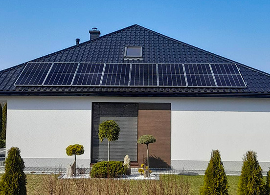 Panele słoneczne na ciemnym dachu nowoczesnego domu jednorodzinnego z białymi ścianami i starannie utrzymanym ogrodem z ozdobnymi drzewkami.