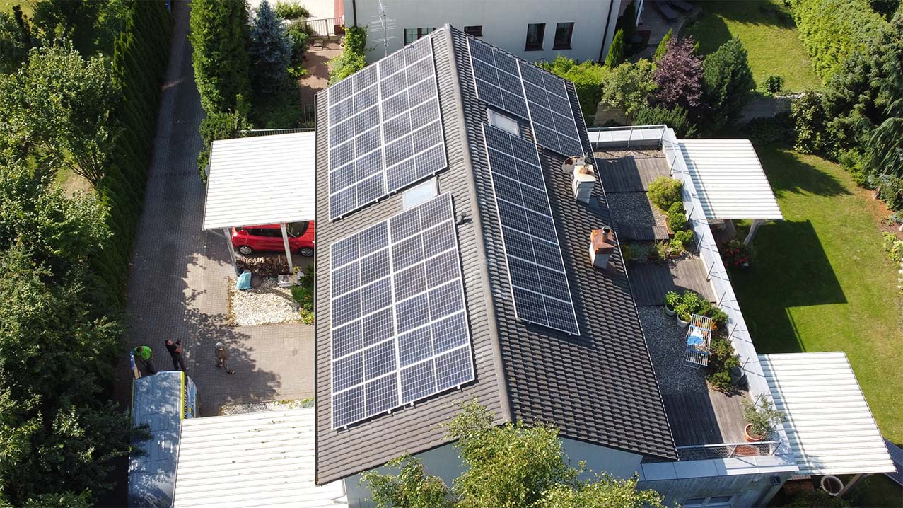 Widok z góry na dom z panelami słonecznymi na dachu, obok taras na dachu z roślinami, czerwony samochód w wiacie, pracownicy przy samochodzie dostawczym.