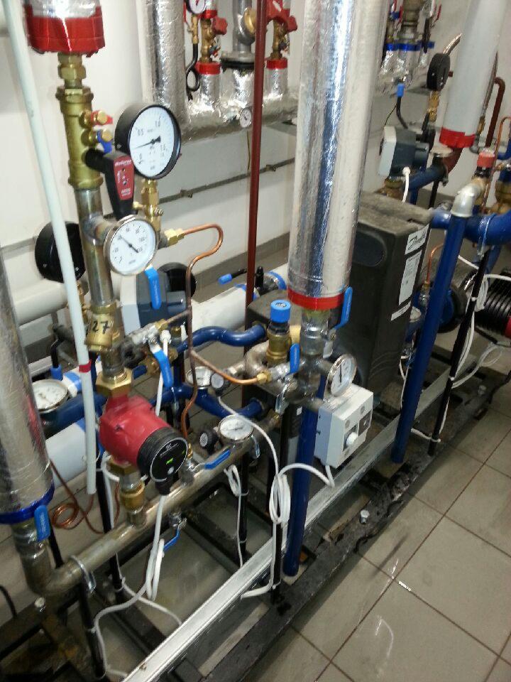 Złożona instalacja z rur, zaworów, manometrów i pompy obiegowej, z widocznymi elementami izolacji termicznej na tle szarej ściany i płytek podłogowych.