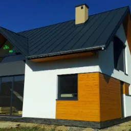 Gotowy dom szkieletowy z ciemną blachodachówką, jasną elewacją z drewnianymi akcentami, dużymi oknami i napisem 'Eko 889' na szczycie