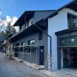 Dwukondygnacyjny dom z szarą elewacją, częściowo ocieplony białą farbą, z rozstawionym rusztowaniem i drabiną przy otwartym garażu, jesienne otoczenie.