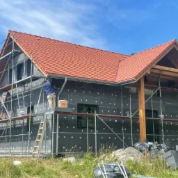 Dom jednorodzinny w trakcie ocieplania: czarne płyty izolacyjne z białymi punktami na elewacji, pracownicy na rusztowaniu, dach z czerwonej dachówki, błękitne niebo.