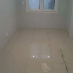 Nowo położone, beżowe płytki podłogowe w pokoju z szarymi ścianami, widoczne dwa białe wiadra i poziomica, okno z dekoracyjną folią.