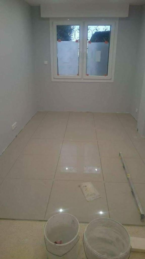 Nowo położone, beżowe płytki podłogowe w pokoju z szarymi ścianami, widoczne dwa białe wiadra i poziomica, okno z dekoracyjną folią.