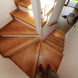 Arpex glasswood - Drewniane schody z białymi barierkami, widok z góry na stopnie o trójkątnym kształcie. Na dole kadru widoczne stopy w skarpetach. W tle odkurzacz.