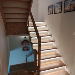 Arpex glasswood - Drewniane schody z podświetleniem LED, widok z dołu. Balustrada z drewna i szkła. Na ścianie wiszą obrazy w czarnych ramkach. Widoczne drewniane wykończenie pod schodami.