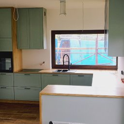 Arpex glasswood - Jasna kuchnia z drewnianymi blatami i zielonymi szafkami, widok na okno z rusztowaniami. Nowoczesny design, minimalistyczne uchwyty, czarna armatura.