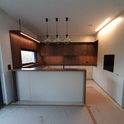 Arpex glasswood - Nowoczesna kuchnia na wymiar z drewnianymi elementami i wyspą, oświetlona wiszącymi lampami. Widoczne niedokończone wykończenie podłogi.