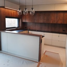 Arpex glasswood - Nowoczesna kuchnia z drewnianymi szafkami i wyspą, oświetlona wiszącymi lampami. Widoczne zabezpieczenia podłogi, sugerujące świeży montaż.