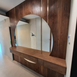 Arpex glasswood - Drewniana zabudowa ściany z owalnym lustrem i szafkami z dekoracyjnymi, złotymi wstawkami. Widoczne detale wykończenia i odbicie wnętrza w lustrze.