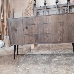 Arpex glasswood - Drewniana komoda z szufladami i szafkami na czarnych nóżkach, wykonana na wymiar w Lędzinach. Widoczne słoje drewna i detale wykończenia. W tle warsztat stolarski.