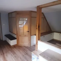 Pokój na poddaszu z drewnianą zabudową: drzwi z przeszkleniami, szafki wnękowe pod skosem, regał na książki i drewniany parkiet.