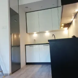 Nowoczesna kuchnia w odcieniach bieli i czerni, z połyskującymi frontami szafek i zabudowaną lodówką, oświetlona punktowymi lampami.