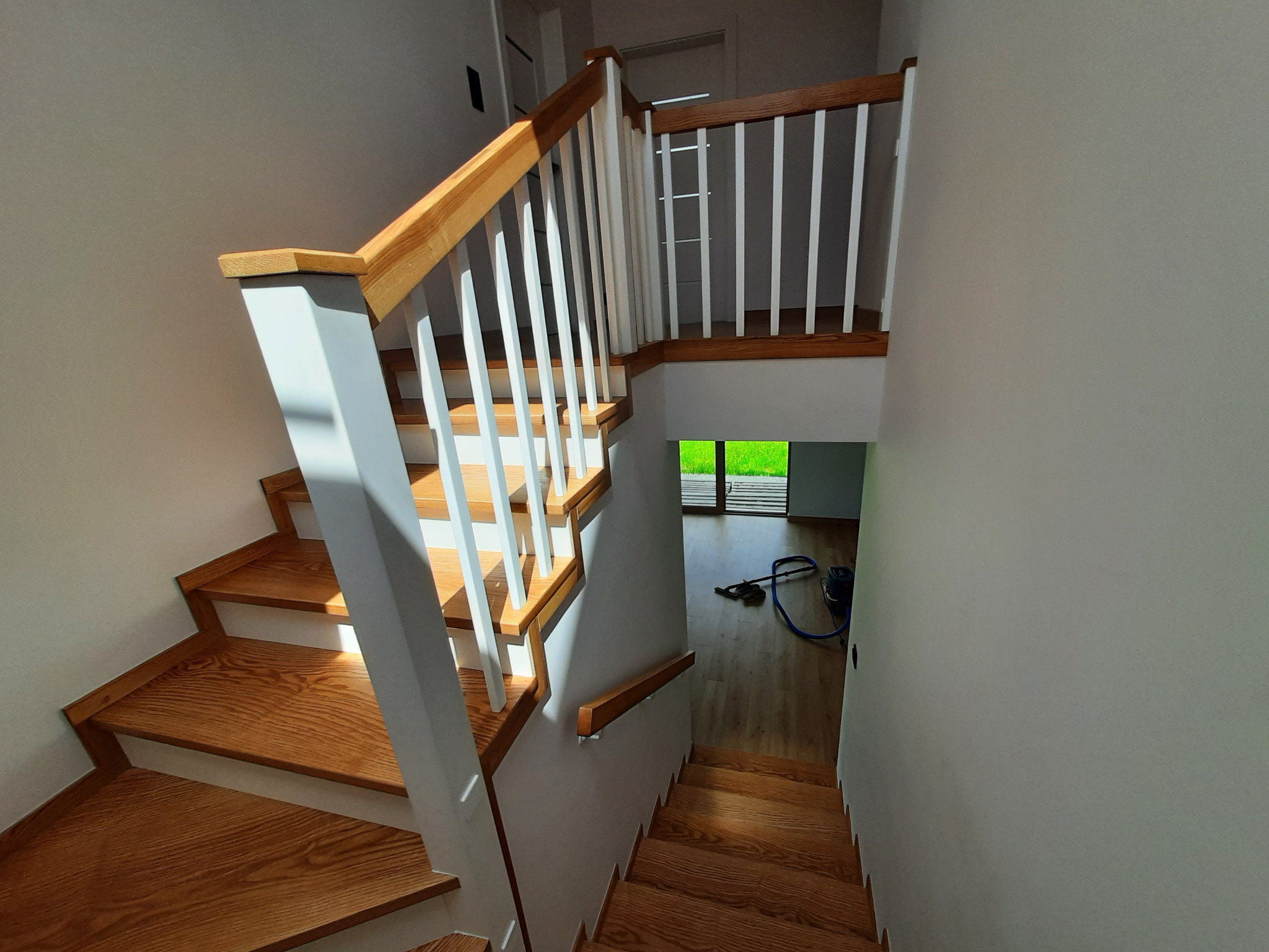 Drewniane schody z białymi balustradami w jasnym wnętrzu. Widok z góry na dół, z widocznym fragmentem podłogi i sprzętem czyszczącym w tle. Naturalne oświetlenie.