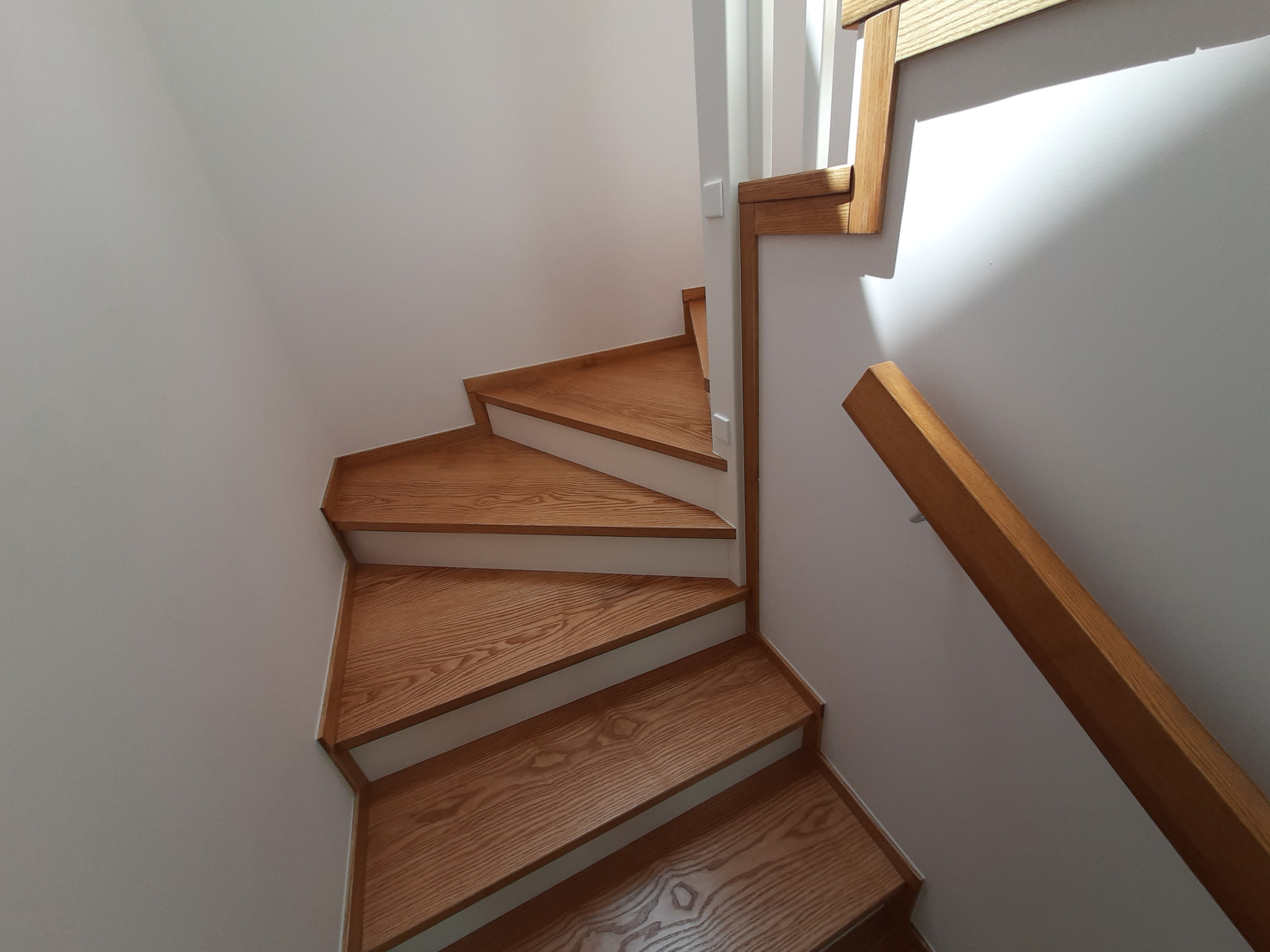 Drewniane schody z białymi podstopnicami i drewnianą balustradą, widziane z góry, oświetlone naturalnym światłem. Detal wykończenia wnętrza.