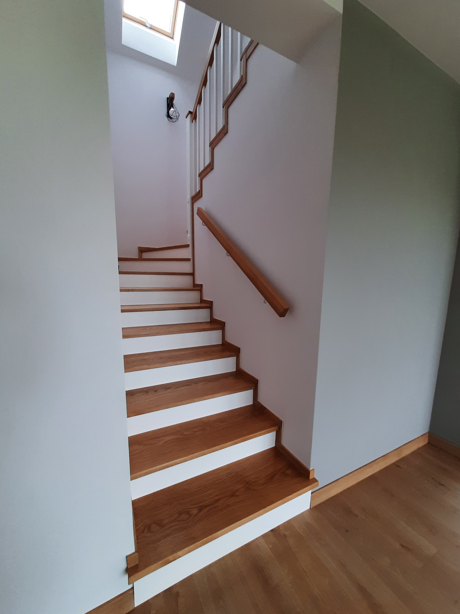 Drewniane schody z białymi podstopnicami w jasnym wnętrzu. Naturalne oświetlenie z okna dachowego. Minimalistyczny design z drewnianą poręczą.