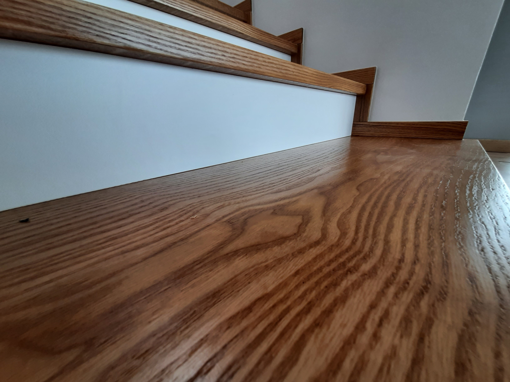 Polerowane drewniane schody z widocznym usłojeniem, jasne stopnice i białe podstopnice, ujęcie z dołu podkreślające fakturę drewna.